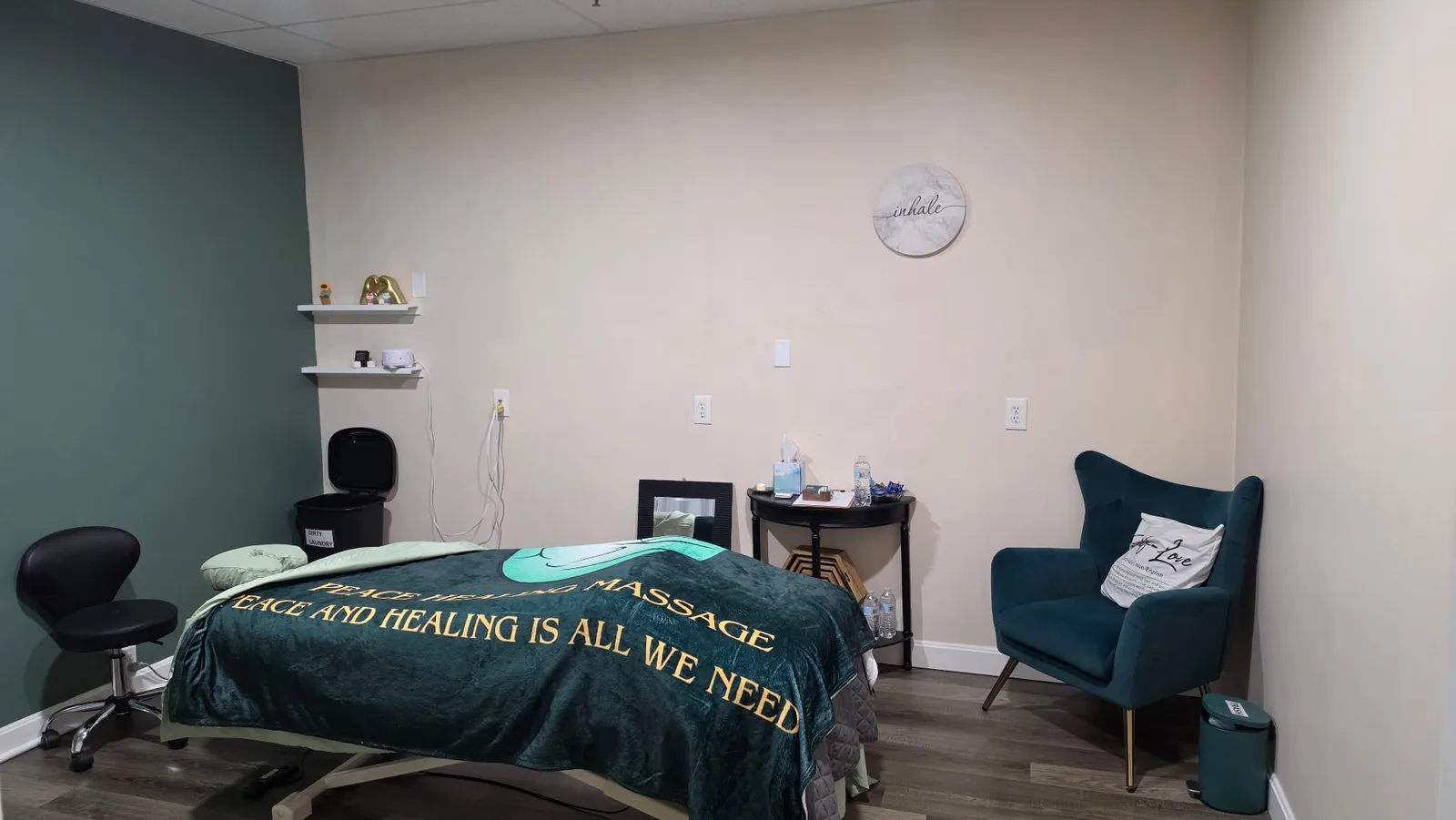 Treatment room at Peace Healing Massage with the massage table dressed in a green Peace Healing Massage blanket, teal armchair and inhale wall art